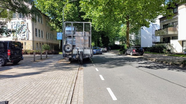 Rückert Straße in Osnabrück. Blickrichtung Schule bzw. Ernst-Sievers-Straße. Auf dem schmalen Schutzstreifen links entgegen der Fahrtrichtung steht ein beladener Anhänger und blockiert eben diesen. Ein Auto fährt am Anhänger vorbei.