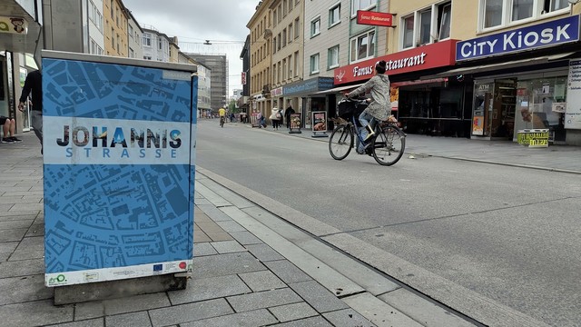 Johannisstraße in Osnabrück. Blickrichtung Neumarkt. Links im Vordergrund ein Mülleimer mit Johannisstraßen Design. Rechts der City Kiosk. Auf der Johannisstraße (Fußgängerzone) eine Radfahrerin.