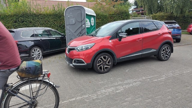 Rotes Auto steht in einer Tempo 30 Zone auf der "Fahrbahn". Auf der Seitenscheibe ist ganz klein etwas Aufgeklebtes zu erkennen. Im Hintergrund sind Parkplätze zu sehen.
