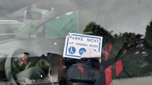 CloseupAufkleber mit dem Text "Parke nicht auf unseren Wegen" auf einer Seitenscheibe eines Autos. Es wurde versucht den Aufkleber zu entfernen. 