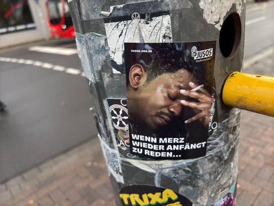 Das Foto zeigt einen Aufkleber an einem Verkehrspoller in einer städtischen Umgebung. Der Aufkleber zeigt eine Person, die sich die Stirn hält und eine Zigarette zwischen den Fingern hat. Oben rechts steht "JUSOS Niedersachsen". Der Text auf dem Aufkleber lautet: "WENN MERZ WIEDER ANFÄNGT ZU REDEN...". Der Poller ist mit weiteren Aufklebern und Bemalungen bedeckt. Im Hintergrund ist eine Straße und ein rotes Fahrzeug zu sehen.