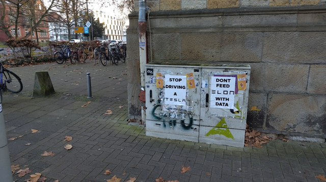 Das Bild zeigt einen grauen Verteilerkasten an einer steinernen Gebäudeecke im Freien. Auf dem Kasten sind mehrere Aufkleber und Graffiti angebracht. Besonders auffällig sind zwei Plakate mit den Aufschriften "Stop Driving a Tesla" und "Don't Feed Elon with Data". Im Hintergrund sind Fahrräder, Bäume und Gebäude zu sehen. Der Boden ist mit Pflastersteinen ausgelegt und mit einigen Herbstblättern bedeckt.