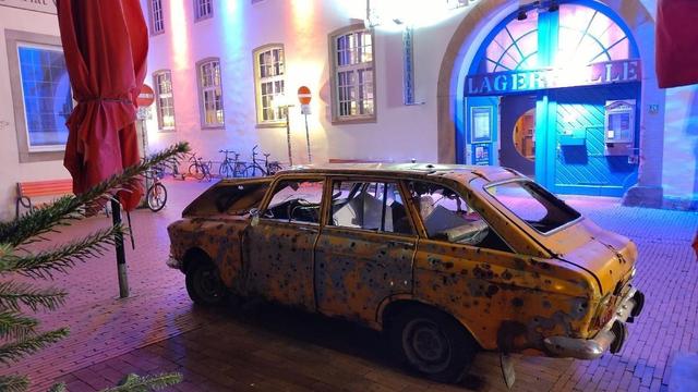 Ein zerschossenes ukrainisches Auto steht als Kunstprojekt vor einem Gebäude mit der Aufschrift "LAGERHALLE".