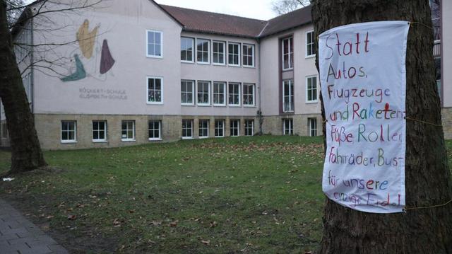 Das Bild zeigt eine Außenaufnahme eines Schulgebäudes, der Rückert-Schule Elisabeth-Schule, vor dem ein Baum steht. An dem Baum ist ein handgemachtes Protestplakat befestigt. Auf dem Plakat steht in bunten Buchstaben die Forderung, statt Autos, Flugzeuge und Raketen lieber Füße, Roller, Fahrräder und Busse zu nutzen, "für unsere einzige Erde". Die Botschaft des Plakats ist ein Aufruf zu umweltfreundlicherer Mobilität und zum Schutz der Erde.