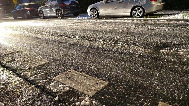 Das Bild zeigt eine Straße, die mit einer Mischung aus Eis und Schnee bedeckt ist. Im Hintergrund sind drei Autos zu sehen, die ebenfalls Spuren von Schnee tragen. Die Straße reflektiert das Licht, was auf glatte und potenziell gefährliche Bedingungen hindeutet. Reifenspuren im Schnee deuten darauf hin, dass Fahrzeuge kürzlich die Straße befahren haben.