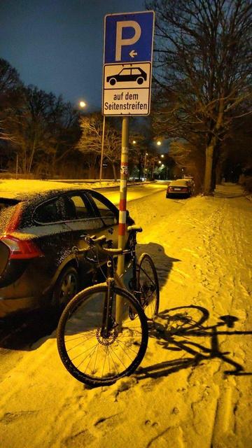 Das Bild zeigt eine verschneite Straßenszene im frühen Sonntagmorgen. Ein Fahrrad ist an einem Pfosten gelehnt, an dem ein blaues Parkplatzschild mit der Aufschrift "auf dem Seitenstreifen" angebracht ist. Im Hintergrund stehen Autos am Straßenrand, und die gelbliche Beleuchtung erzeugt lange Schatten im Schnee.