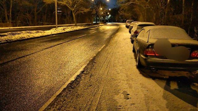 Das Bild zeigt eine Straße im frühen Sonntag Morgen mit einer dicken Eisschicht auf dem Fahrrad Schutzstreifen. Zwei Fahrspuren stadtauswärts sind akkurat frei/geräumt. Entlang der Straßenseite stehen geparkte Autos, und die Straße ist nass und reflektiert das Licht der nahegelegenen Straßenlaternen.