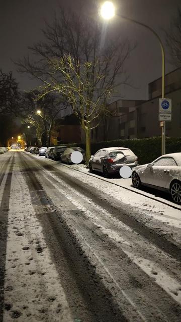 Die Aufnahme zeigt eine verschneite Straße im frühen Morgen, mit parkenden Autos und Straßenlaternen. Es ist noch dunkel. Schnee bedeckt Autos und die Fahrbahn. Es ist Winter. Am rechten Fahrbahnrand steht ein Schild "Fahrradstraße". Auf der Fahrbahn ist bruchstückhaft auch "Fahrradstraße" zu erahnen.