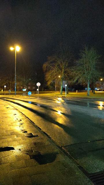 Das Bild zeigt eine morgendliche Aufnahme einer Straße mit Straßenlaternen. Die Straße ist nass, was zu Reflexionen des Lichts führt. Am Straßenrand stehen Bäume und Verkehrszeichen. Der Himmel ist dunkel.