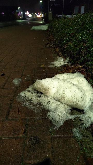 Das Bild zeigt am frühen Morgen einen Radweg mit Pflastersteinen. Am rechten Bildrand befindet sich ein Grünstreifen mit Büschen und Bäumen. Auf dem Gehweg liegt schmelzender Schnee, der teilweise mit Schmutz bedeckt ist. Im Hintergrund sind Straßenlaternen und ein unscharfes Licht von Autos zu sehen. Die Szenerie wirkt ruhig und etwas verlassen.
