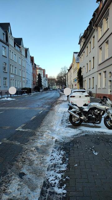 Das Bild zeigt eine Straße in einer Stadt an einem Wintertag. Entlang der Straße stehen mehrgeschossige Gebäude mit unterschiedlicher Architektur. Am Straßenrand liegt Schnee, der teilweise schon geschmolzen ist. Ein Motorrad steht geparkt am rechten Straßenrand. Im Hintergrund sind weitere Autos und Bäume zu sehen. Der Himmel ist klar und blau.
