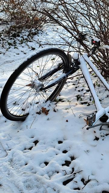 Das Bild zeigt ein umgekipptes Fahrrad, das teilweise im Schnee liegt. Der Rahmen des Fahrrads ist mit Schnee bedeckt, und die Umgebung ist ebenfalls mit Schnee bedeckt. Im Hintergrund sind Büsche oder kleine Bäume zu sehen. Das Bild vermittelt eine winterliche und ruhige Atmosphäre.