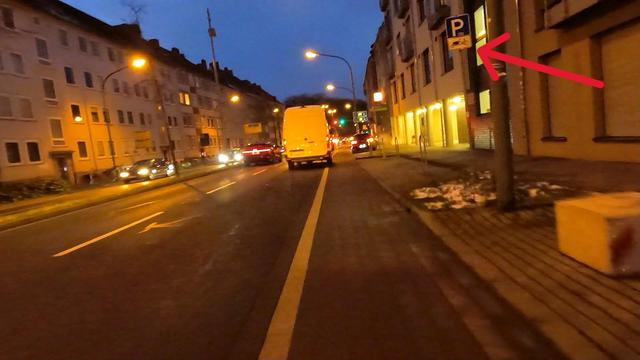 Das Bild zeigt eine Straße, den Johannistorwall in Osnabrück bei Dämmerung. Auf der rechten Seite ist ein gelber Paketlieferwagen auf der Fahrbahn für Autos geparkt. Es gibt zwei Fahrspuren. Der Schutzstreifen für Radfahrer und Parkplätze für Lastenräder daneben bleiben frei. Links und rechts im Bild sind mehrere mehrstöckige Gebäude zu sehen, mit beleuchteten Fenstern. Straßenlaternen erhellen die Szene, und es sind vereinzelt Autos zu sehen. 