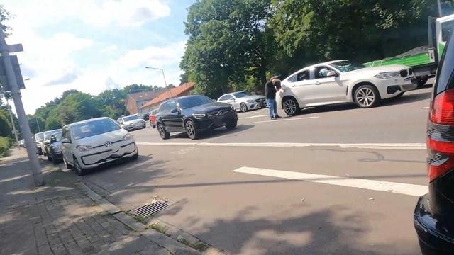 Auf dem Bild sieht man eine städtische Straße bei Tageslicht. Mehrere Autos stehen oder fahren im Verkehr, einige sind am Straßenrand geparkt. In der Mitte der Straße stehen zwei Autos dicht beieinander, daneben eine Person auf der Fahrbahn, die einen weißen SUV mit einem 5l Kanister betankt. Am Rand sieht man einen Gehweg, Bäume mit viel Grün und Häuser im Hintergrund.