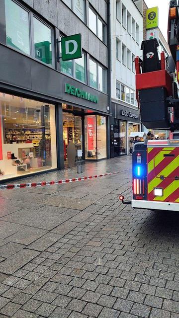 Auf dem Bild sieht man eine Fußgängerzone mit mehreren Geschäften. Im Mittelpunkt ist ein Deichmann-Schuhladen mit großen Schaufenstern. Der Bereich davor ist mit rot-weißem Absperrband der Polizei abgesperrt. Rechts steht ein großes Feuerwehrfahrzeug mit ausgefahrenem Hubarm und eingeschaltetem Blaulicht. Der Boden ist nass mit Schneematsch, es wirkt wie nach Regen, und einige Passanten stehen weiter hinten und schauen zu.