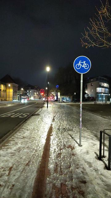 Das Foto zeigt eine nächtliche Straßenszene im Winter in Osnabrück. Ein Geh- und Radweg an der Straße "Neuere Graben" am Ledenhof ist mit Eis und festgetretenem Schnee bedeckt und wirkt glatt und glänzend im Licht der Straßenlampen. Rechts im Vordergrund steht ein blaues rundes Verkehrsschild mit einem weißen Fahrradsymbol, das einen Radweg kennzeichnet.

Links daneben verläuft eine Straße mit einer markierten Busspur; Autos mit eingeschalteten Scheinwerfern fahren in der Ferne. Mehrere Straßen…