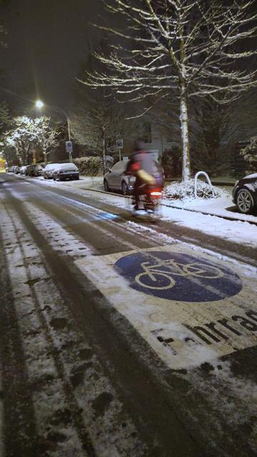 Das Bild zeigt eine morgendliche Straßenszene im Winter. Es liegt Schnee auf der Fahrbahn, den Gehwegen und auf parkenden Autos. Die Straße ist von Laternen beleuchtet, deren Licht sich gelblich im Schnee spiegelt.

Im Vordergrund ist deutlich ein blaues Fahrradsymbol auf der Fahrbahn zu sehen – ein markierter Radweg. Daneben steht teilweise lesbar das Wort „Fahrrad…“ (vermutlich „Fahrradstraße“ oder ein Hinweis auf den Radverkehr). Der Schnee ist von Reifenspuren durchzogen.

In der Mitte des …