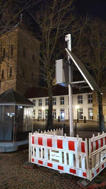 Auf demm nächtlichen Theatervorplatz steht ein abgesperrter Mast mit Technik. Der Mast ist von rot-weißen Baustellenabsperrungen umgeben. Oben daran sind ein schräg stehendes Solarpanel, eine Kamera oder ein Sensor sowie ein grauer Technik-Kasten montiert.

Im Hintergrund sieht man ein beleuchtetes Gebäude mit mehreren Fenstern und der Osnabrücker Dom. Rundherum stehen kahle Bäume, und der Boden ist mit Kopfsteinpflaster bedeckt. 🌙

