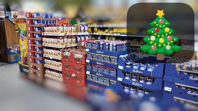 Hier ist eine kurze Bildbeschreibung:
Das Bild zeigt das Innere eines Supermarktes aus einer leicht erhöhten Perspektive. Links stehen hohe Stapel roter Kartons, daneben Regale mit weiß verpackten Ware. Im Detail Weihnachtsgebäck. Dahinter befinden sich weitere Regale unter hellem Licht. Auf der rechten Seite ist ein digitaler Weihnachtsbaum eingeblendet – grün, mit bunten Kugeln und gelbem Stern. Das Bild hat einen weichen Fokus an den Rändern. Das ganze im Sommer August 2026.